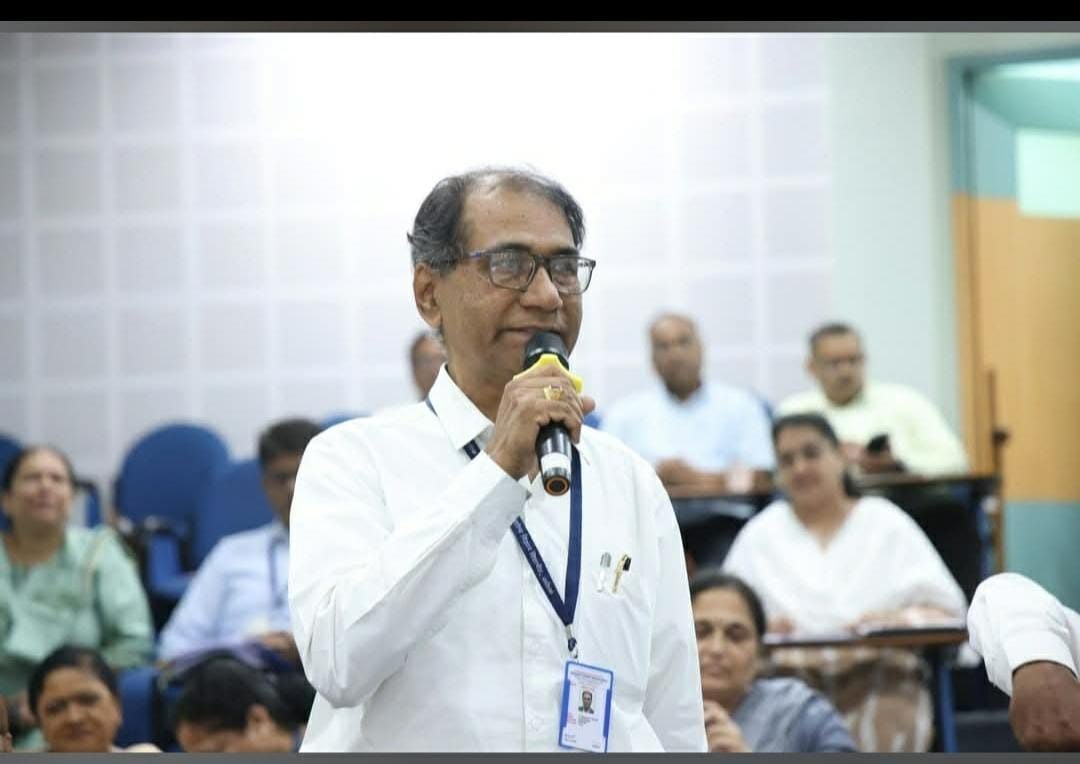 A speaker at a microphone addressing a meeting, with seated participants in the room.